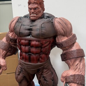 May include: A large, muscular figurine of a character with red hair and a stern expression. The figure is predominantly red and brown, with detailed musculature and armor-like elements. The figure's arms are massive, and it has a determined facial expression.
