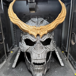 May include: A 3D printed skull-shaped helmet with a metallic silver finish and a textured surface. The helmet features large, curved, gold-colored horns extending from the forehead and a detailed skeletal face with deep eye sockets and exposed teeth.