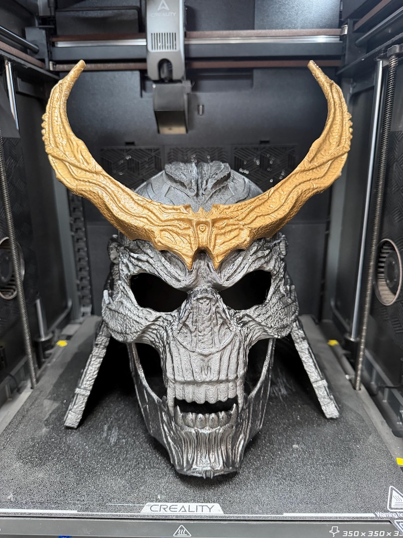 May include: A 3D printed skull-shaped helmet with a metallic silver finish and a textured surface. The helmet features large, curved, gold-colored horns extending from the forehead and a detailed skeletal face with deep eye sockets and exposed teeth.
