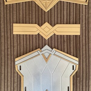 May include: Three gold-coloured plastic pieces for a Wonder Woman costume. The top piece is a headband with a star in the centre. The middle piece is a belt with a geometric design. The bottom piece is a chest piece with a silver centre and a gold border.