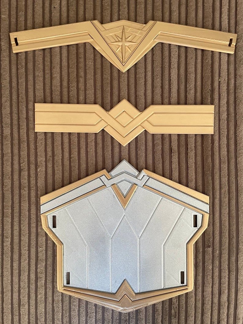 May include: Three gold-coloured plastic pieces for a Wonder Woman costume. The top piece is a headband with a star in the centre. The middle piece is a belt with a geometric design. The bottom piece is a chest piece with a silver centre and a gold border.