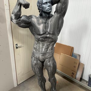 May include: A gray statue of a bodybuilder flexing his muscles. The statue is detailed with realistic muscle definition and is posed in a classic bodybuilding stance. The statue is standing in front of a white door.
