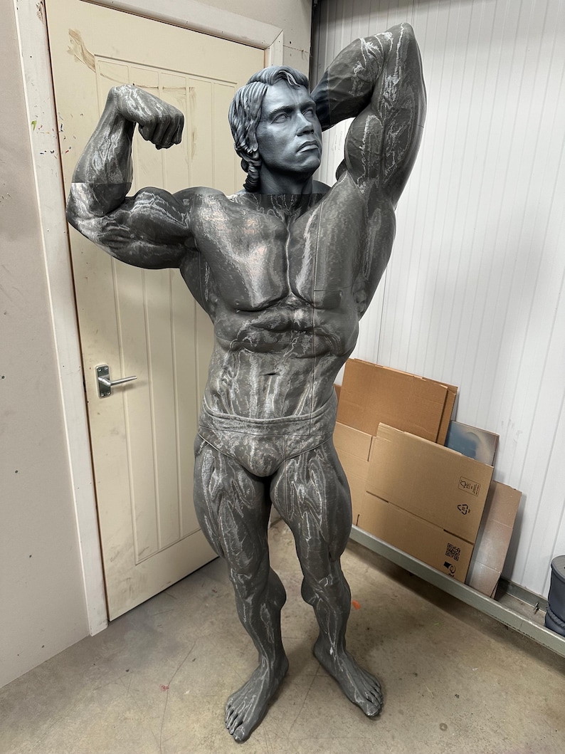 May include: A gray statue of a bodybuilder flexing his muscles. The statue is detailed with realistic muscle definition and is posed in a classic bodybuilding stance. The statue is standing in front of a white door.