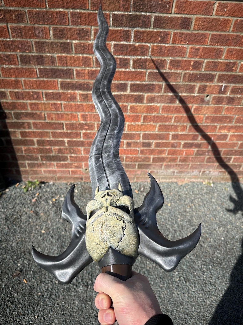 May include: A fantasy sword with a curved, silver blade and a skull-shaped hilt. The hilt is tan and brown, with two curved, black wings extending from the sides. The sword has a long, wavy blade.