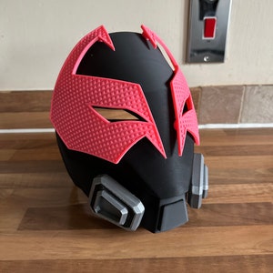 May include: A black and pink 3D printed mask with a geometric design. The mask has a black base with a pink geometric pattern on the front. The mask has two silver-colored accents on the sides.