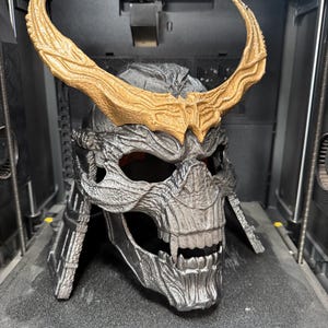 May include: A 3D printed skull-shaped helmet with a metallic silver finish and prominent fangs. The helmet features large, textured, gold-colored horns extending from the forehead. The design includes intricate, weathered detailing across the surface.