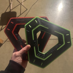 May include: Two geometric, hexagonal-shaped objects. One is black with a red outline, and the other is black with a green outline. The objects have a textured surface and are held in a hand.