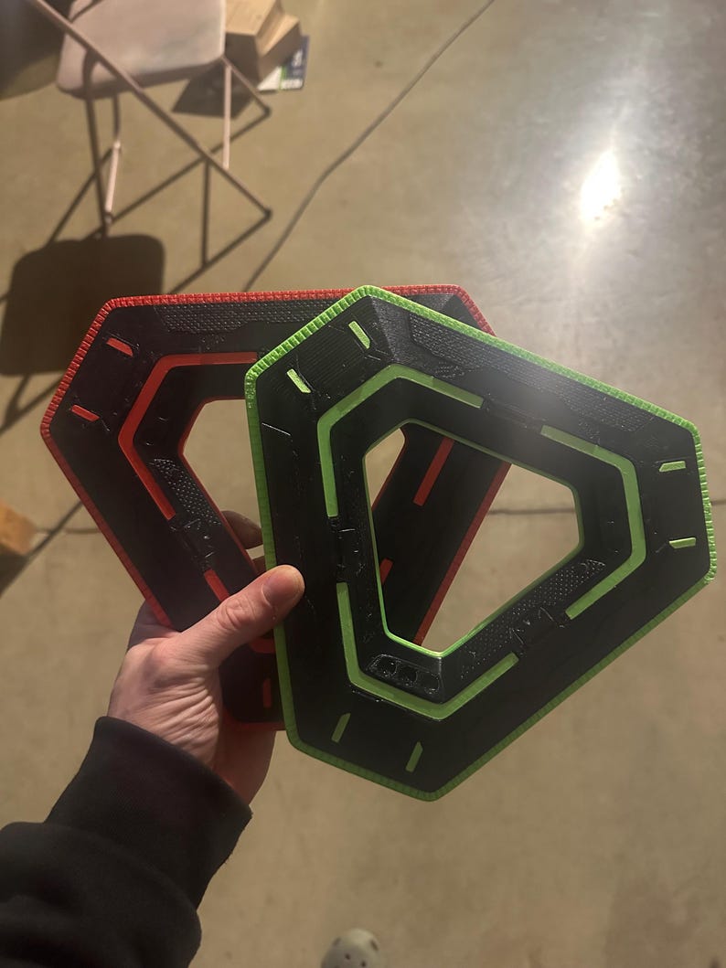 May include: Two geometric, hexagonal-shaped objects. One is black with a red outline, and the other is black with a green outline. The objects have a textured surface and are held in a hand.