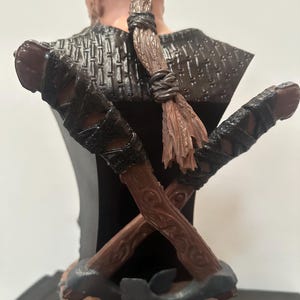 May include: A detailed bust of a warrior with a braided hairstyle and crossed axes. The bust features a dark colour palette with brown, black, and grey tones. The axes are intricately detailed with wooden handles and leather wrappings, and the base has Celtic knot designs.