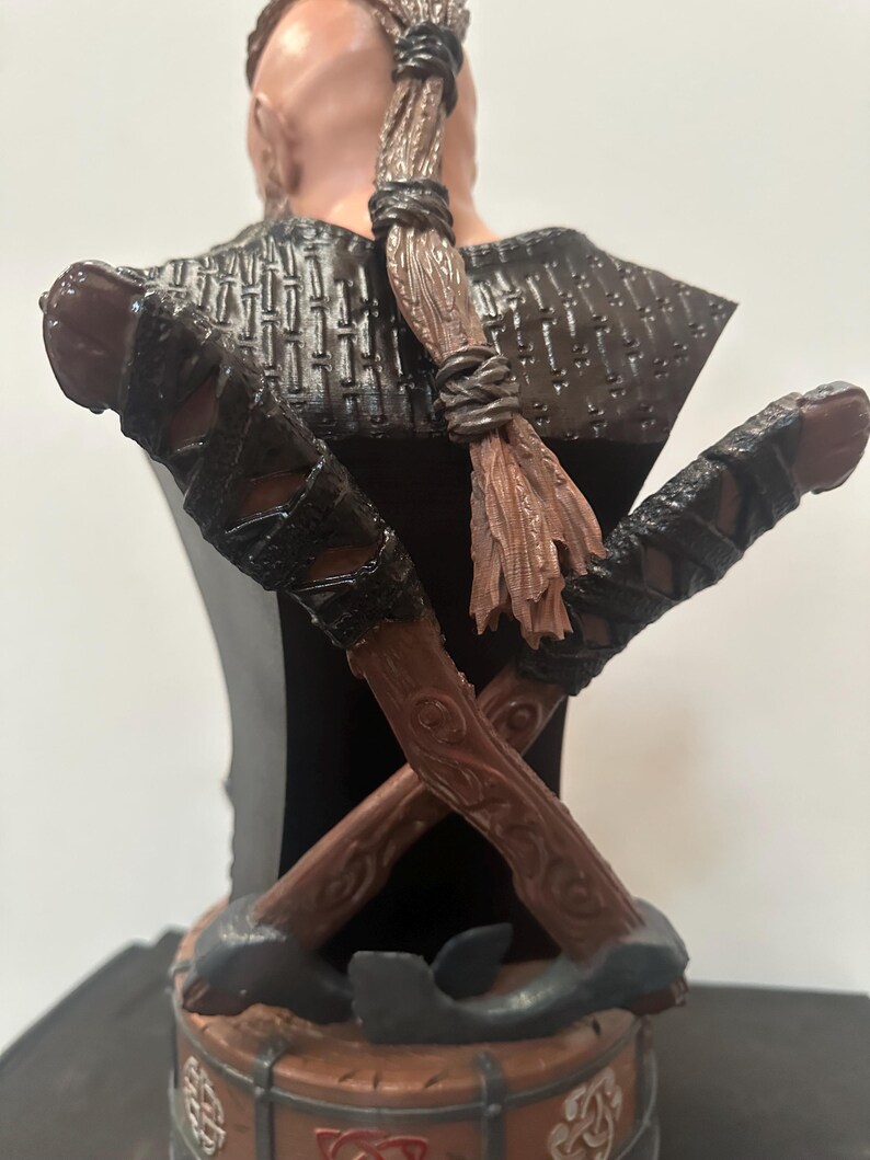 May include: A detailed bust of a warrior with a braided hairstyle and crossed axes. The bust features a dark colour palette with brown, black, and grey tones. The axes are intricately detailed with wooden handles and leather wrappings, and the base has Celtic knot designs.