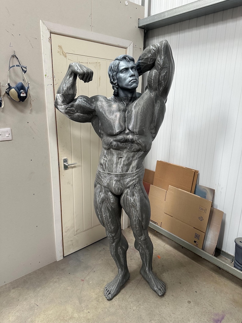 May include: A gray statue of a bodybuilder in a classic pose, flexing his biceps. The statue is detailed with realistic muscle definition and is wearing briefs. The figure is standing in a workshop setting.