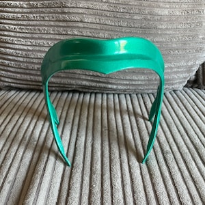 K&ouml;nnte beinhalten: Ein gr&uuml;nes Stirnband mit einem geschwungenen Design. Das Stirnband besteht aus einem glatten, gl&auml;nzenden Material.