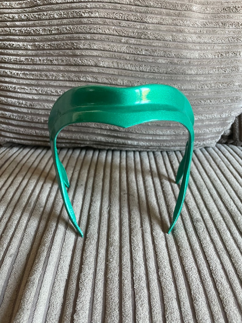 K&ouml;nnte beinhalten: Ein gr&uuml;nes Stirnband mit einem geschwungenen Design. Das Stirnband besteht aus einem glatten, gl&auml;nzenden Material.