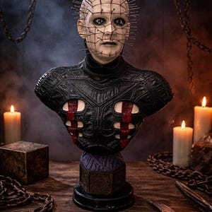May include: A detailed bust of a character with a pale complexion, adorned with metal pins and a grid-like pattern. The figure wears a black leather-like garment with red accents. The bust sits on a decorative base, with chains and candles in the background.