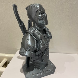 May include: A gray 3D printed bust of Deadpool, a superhero character, with a katana sword on his back. The bust is on a gray base.