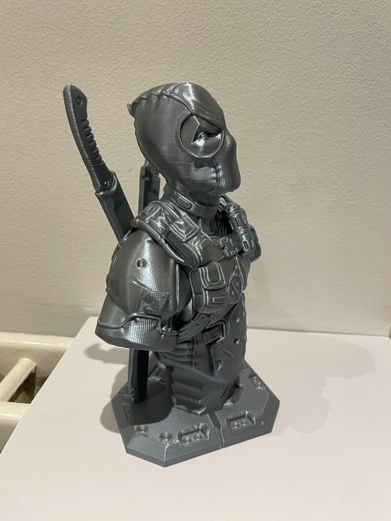 May include: A gray 3D printed bust of Deadpool, a superhero character, with a katana sword on his back. The bust is on a gray base.