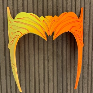 May include: A yellow and orange plastic headband with a decorative design. The headband is curved and has a pointed shape at the top.