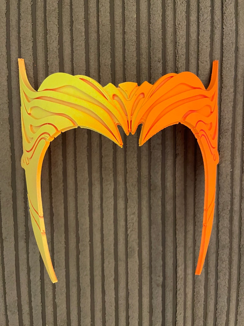 May include: A yellow and orange plastic headband with a decorative design. The headband is curved and has a pointed shape at the top.
