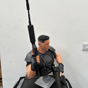 May include: A detailed bust of a character in a black tactical outfit, holding two black rifles. The figure has a determined expression and short, dark hair. The bust is mounted on a gray stand, with a black base.
