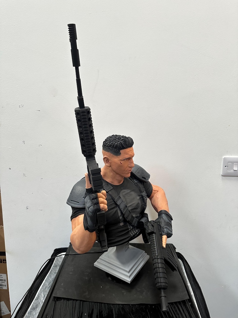 May include: A detailed bust of a character in a black tactical outfit, holding two black rifles. The figure has a determined expression and short, dark hair. The bust is mounted on a gray stand, with a black base.