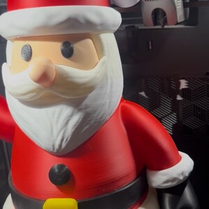 May include: A 3D-printed Santa Claus figure. The figure is predominantly red with white trim on the hat and beard. It has a black belt with a yellow buckle. The figure is a festive decoration.