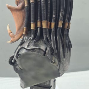 Puede incluir: Busto detallado de un alien&iacute;gena Predator, con un casco y rostro texturizados en tonos grises, mand&iacute;bulas abiertas y un colmillo prominente. Las rastas son negras con acentos beige. El busto est&aacute; montado sobre un pedestal gris texturizado.