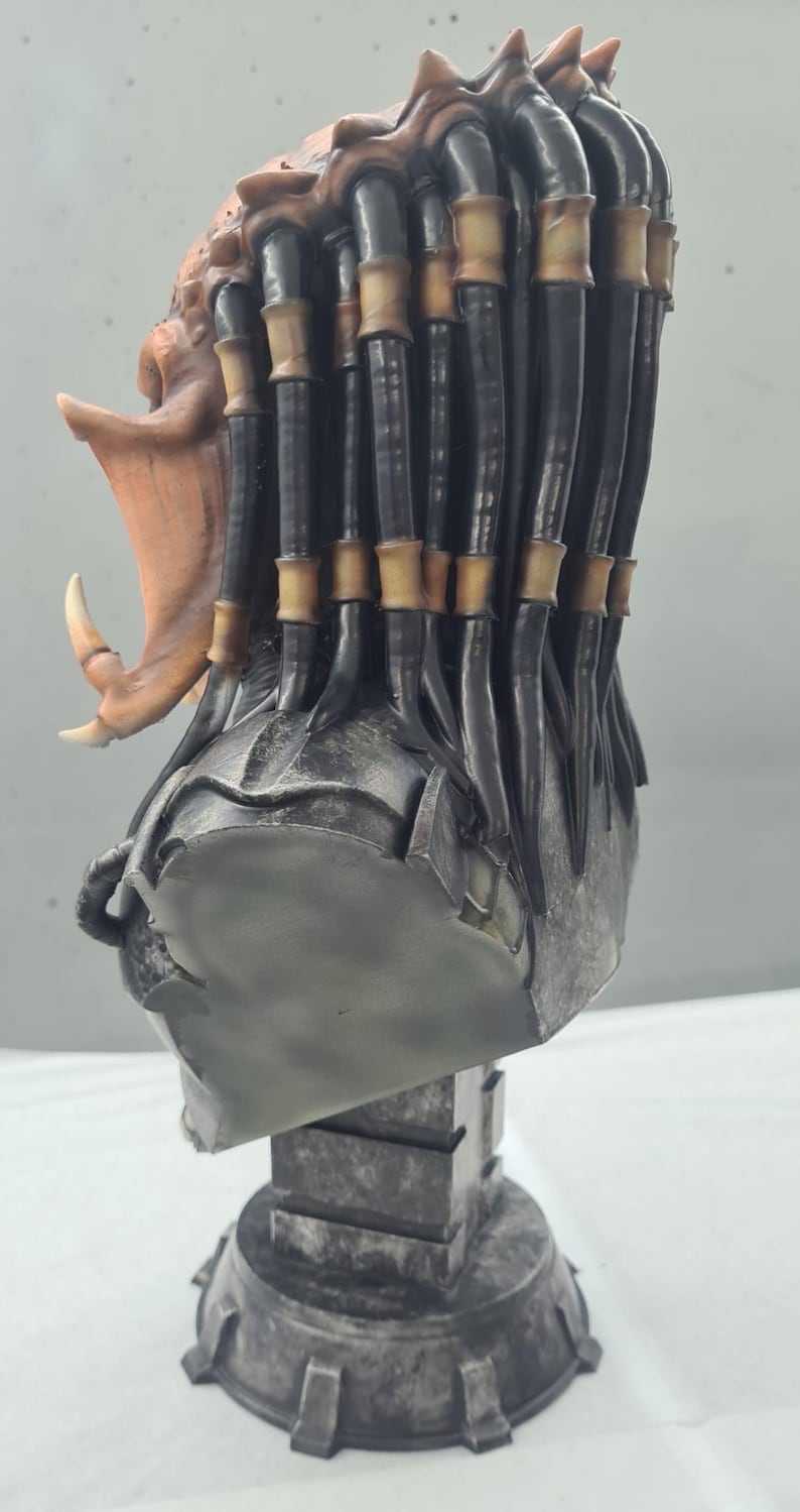 Puede incluir: Busto detallado de un alien&iacute;gena Predator, con un casco y rostro texturizados en tonos grises, mand&iacute;bulas abiertas y un colmillo prominente. Las rastas son negras con acentos beige. El busto est&aacute; montado sobre un pedestal gris texturizado.