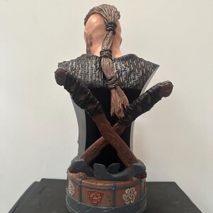 May include: A detailed bust of a Viking warrior, viewed from the back. The figure has a braided hairstyle and is adorned with crossed axes. The base features Celtic knot designs in red and white, with a grey and brown colour scheme.