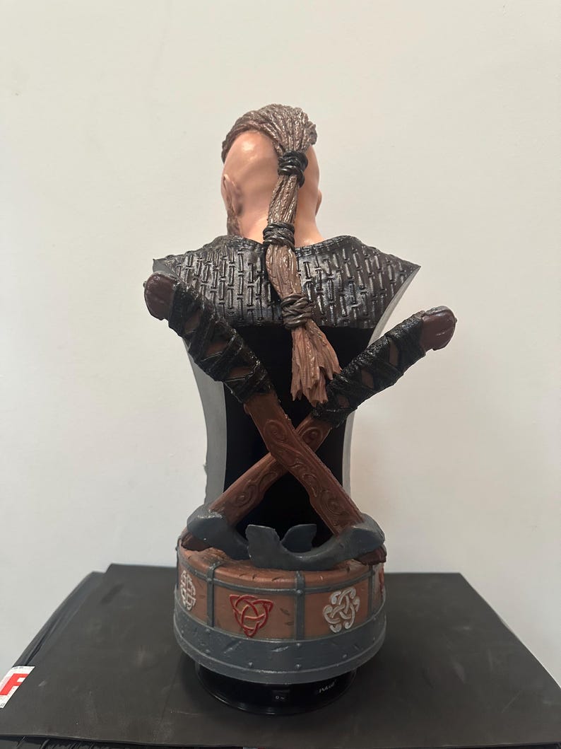 May include: A detailed bust of a Viking warrior, viewed from the back. The figure has a braided hairstyle and is adorned with crossed axes. The base features Celtic knot designs in red and white, with a grey and brown colour scheme.