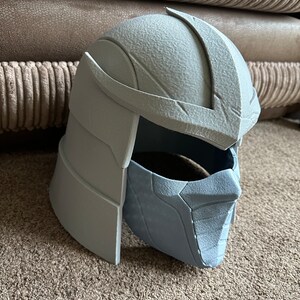 May include: A gray plastic 3D printed helmet with a textured surface. The helmet has a face mask with a detailed design.