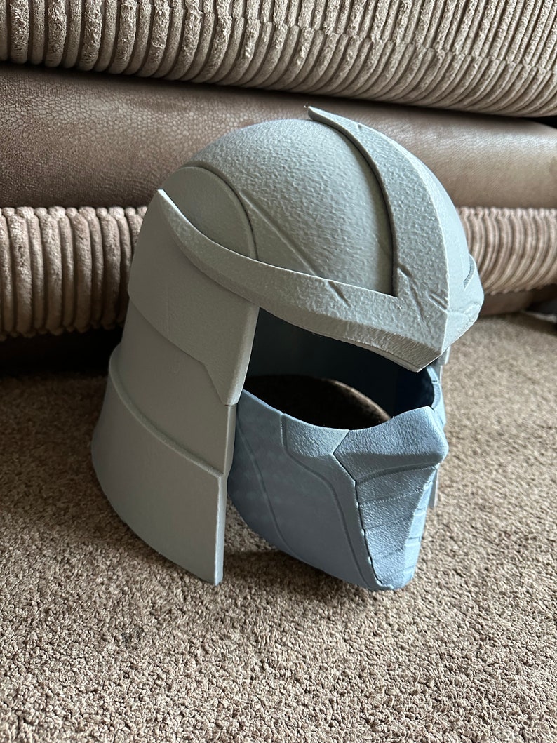 May include: A gray plastic 3D printed helmet with a textured surface. The helmet has a face mask with a detailed design.