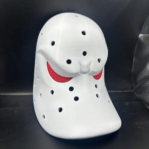 May include: A white mask with multiple small, evenly spaced holes. The mask has a rounded shape with a slightly protruding lower section. Red mesh is visible through the eye openings. The mask is likely a costume or prop.