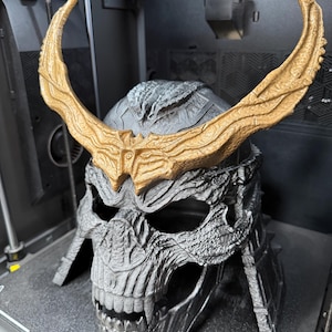 May include: A silver-colored skull-shaped helmet featuring a textured, weathered surface and prominent, large, curved gold-colored horns attached to the forehead. The helmet has deep eye sockets, a defined nasal cavity, and exposed teeth.