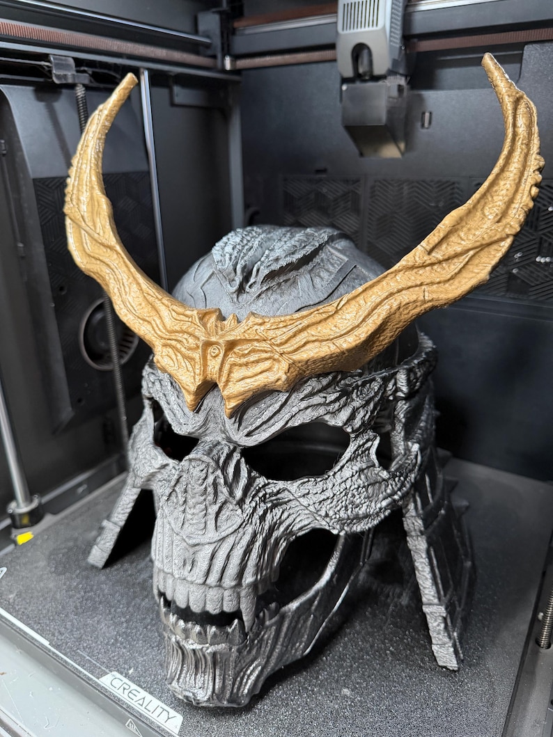 May include: A silver-colored skull-shaped helmet featuring a textured, weathered surface and prominent, large, curved gold-colored horns attached to the forehead. The helmet has deep eye sockets, a defined nasal cavity, and exposed teeth.