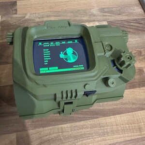 May include: A green 3D printed replica of a Pip-Boy from the Fallout video game series. The screen displays the game's status and special perks menu. The Pip-Boy has a small dial on the side.