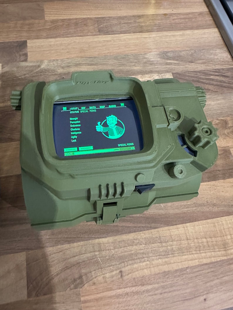 May include: A green 3D printed replica of a Pip-Boy from the Fallout video game series. The screen displays the game's status and special perks menu. The Pip-Boy has a small dial on the side.