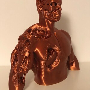 May include: A 3D printed bust of a robot with a detailed, exposed skeletal structure. The bust is made of a copper-colored material and is posed with the head turned to the side.