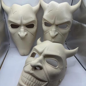 May include: Three off-white, horned masks with menacing expressions. The masks have eye and nose openings, and one features a wide, unsettling grin. The masks are likely for costume or theatrical use.