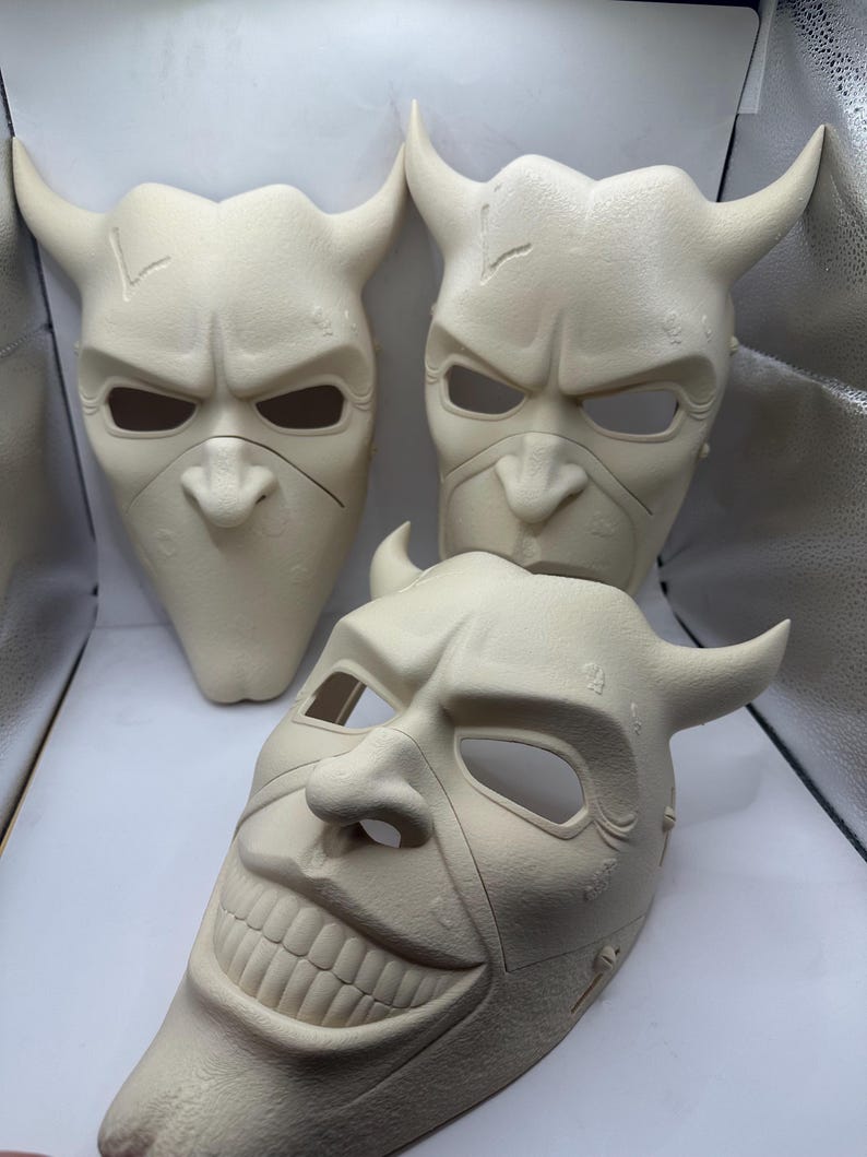 May include: Three off-white, horned masks with menacing expressions. The masks have eye and nose openings, and one features a wide, unsettling grin. The masks are likely for costume or theatrical use.
