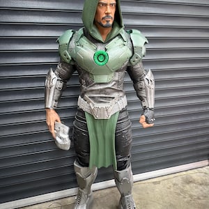May include: A detailed action figure of a character in a green hooded cloak and armor. The figure has a metallic silver and green color scheme, with a green chest piece and a silver belt. The figure is holding a silver mask.