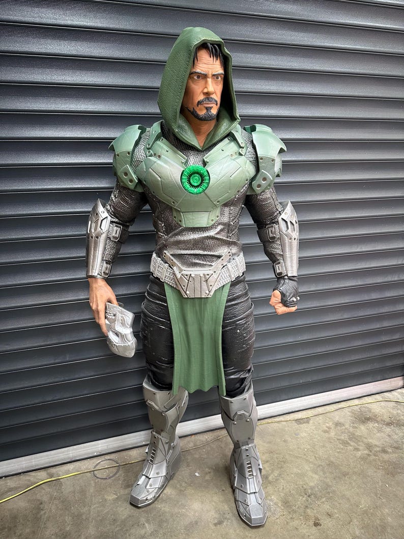 May include: A detailed action figure of a character in a green hooded cloak and armor. The figure has a metallic silver and green color scheme, with a green chest piece and a silver belt. The figure is holding a silver mask.