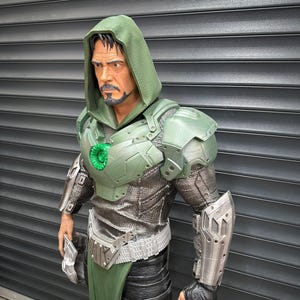 May include: A detailed figurine of a character in a green hooded cloak and armor. The figure has a stern expression and a goatee. The armor is a mix of green and silver, with intricate detailing. The figure is posed with a weapon in one hand.