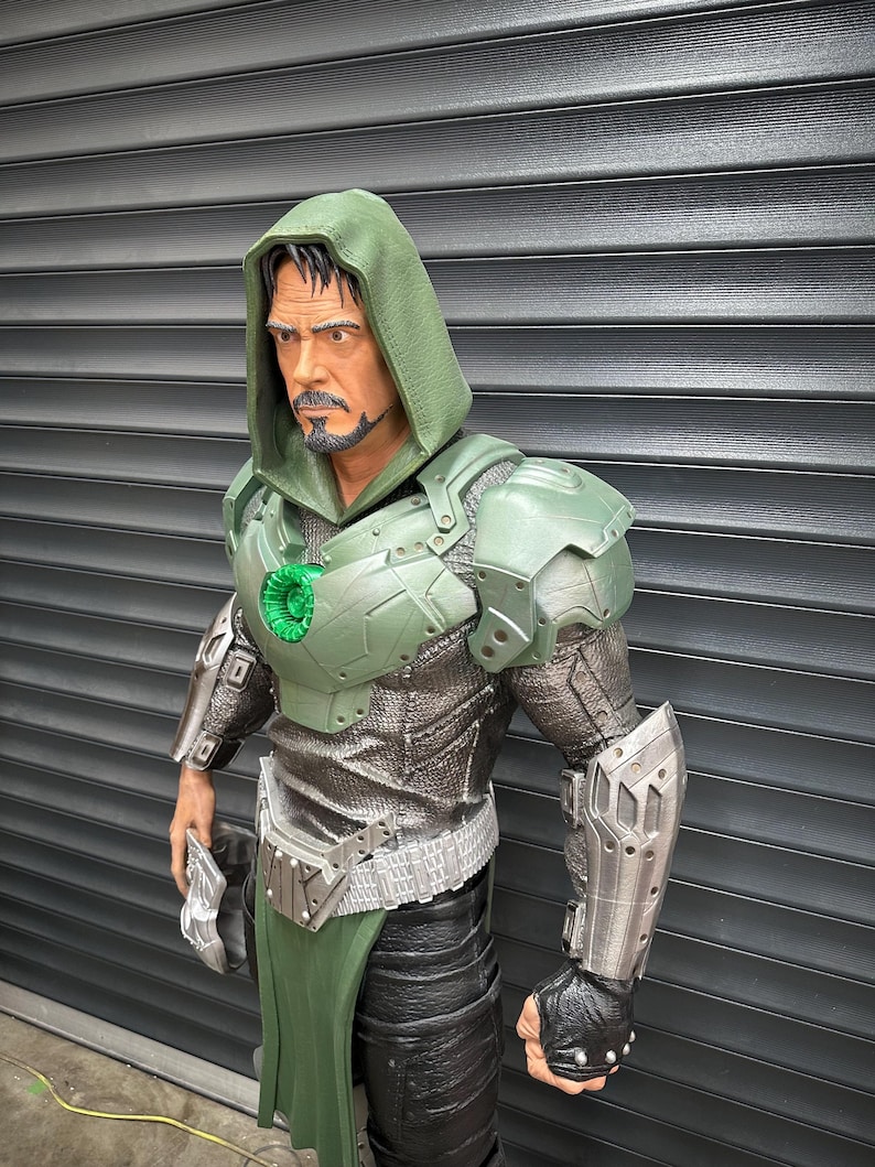 May include: A detailed figurine of a character in a green hooded cloak and armor. The figure has a stern expression and a goatee. The armor is a mix of green and silver, with intricate detailing. The figure is posed with a weapon in one hand.