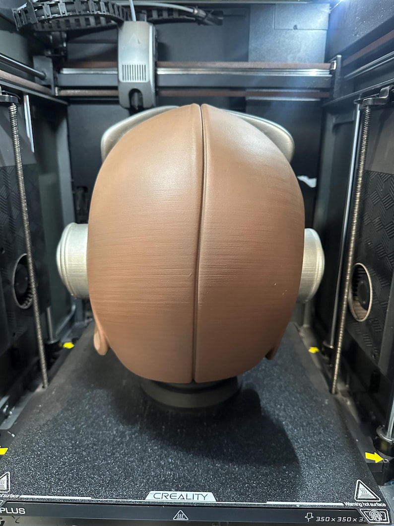 May include: A brown, spherical object is being 3D printed inside a black and silver machine. The object is split into two halves and has a textured surface. The printer bed has the word "CREALITY" printed on it.