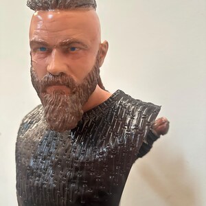 May include: A detailed bust of a man with a full beard and a distinctive hairstyle. The sculpture features a textured, dark-coloured top and is set against a plain background. The man has blue eyes and a serious expression.