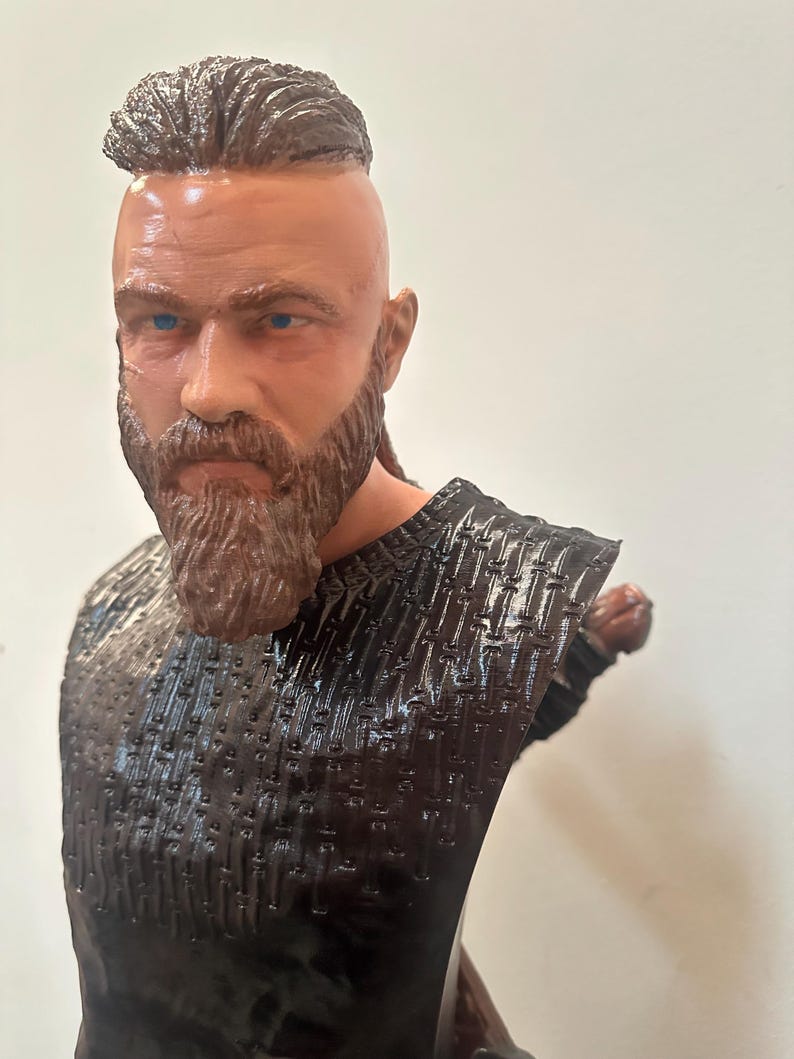 May include: A detailed bust of a man with a full beard and a distinctive hairstyle. The sculpture features a textured, dark-coloured top and is set against a plain background. The man has blue eyes and a serious expression.