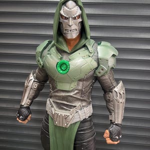 May include: A detailed action figure of Doctor Doom, featuring a silver mask and green hooded cloak. The figure has green armor, a silver chest piece, and black gloves. The figure is posed against a gray background.