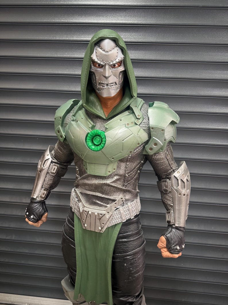 May include: A detailed action figure of Doctor Doom, featuring a silver mask and green hooded cloak. The figure has green armor, a silver chest piece, and black gloves. The figure is posed against a gray background.