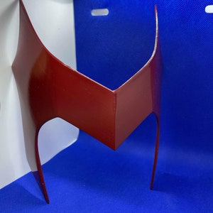 May include: A glossy, maroon-colored headpiece with a distinctive V-shaped design. The headpiece features two elongated, curved side pieces extending downwards. The object is likely a costume accessory or cosplay item.