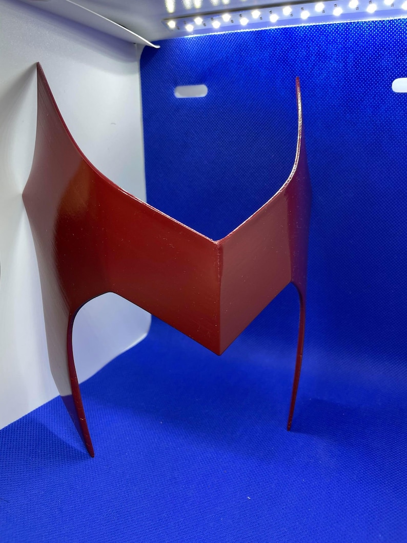 May include: A glossy, maroon-colored headpiece with a distinctive V-shaped design. The headpiece features two elongated, curved side pieces extending downwards. The object is likely a costume accessory or cosplay item.